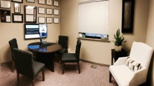 Patient consult area with round black table, 4 black cushioned chair and 1 cream, awards on the wall, and window with blinds pulled
